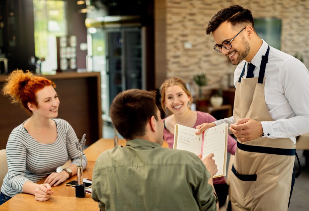 Happy waiter showing menu to a guest who came with friends in a cafe.